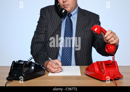 Photo d'un homme assis à un bureau avec deux téléphones traditionnels, un rouge et un noir. Banque D'Images