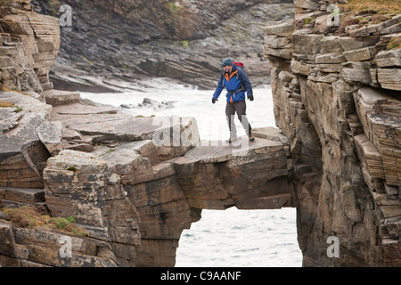 L'alpiniste traverse un pont de pierre cale qui relie les falaises d'une pile de la mer à Yesnaby sur la côte ouest des Orcades Banque D'Images