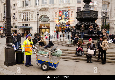 Un nettoyeur de rue et son garrot devant des gens assis sur les marches de la statue d'Eros de Piccadilly Circus London UK Banque D'Images