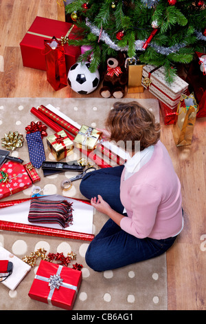 Photo prise à la verticale d'une femme assis sur un tapis à la maison son emballage des cadeaux de Noël. La peluche est générique et n'est pas une marque. Banque D'Images