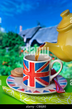 LE CONCEPT DE LA STAYCATION DU COTTAGE AVEC THÉ ANGLAIS. Tasse de thé et biscuits à thème Union Jack anglais, maison typique de chaume et jardin arrière-plan Royaume-Uni Banque D'Images