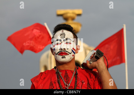 Manifestant une chemise rouge avec visage peint, se tient devant le Monument de la démocratie l'flanqué de drapeaux rouges au coeur de Bangkok. Banque D'Images