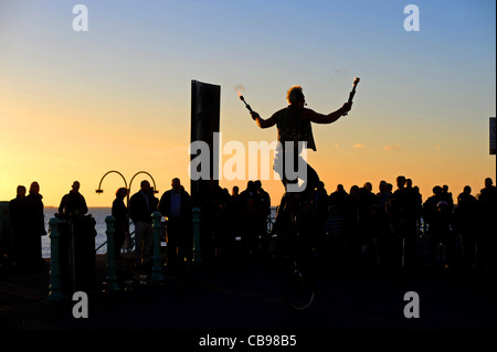 Regarder la foule une flamme juggling musicien ambulant sur un monocycle le long front de mer de Brighton pendant l'temps d'automne exceptionnellement doux Banque D'Images