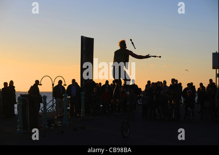 Regarder la foule une flamme juggling musicien ambulant sur un monocycle le long front de mer de Brighton pendant l'temps d'automne exceptionnellement doux Banque D'Images