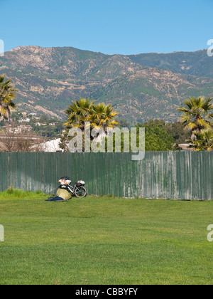 Une personne sans-abri's location s'appuie contre une clôture avec des paquets de biens dans un parc de la ville de Santa Barbara, Californie, USA. Banque D'Images