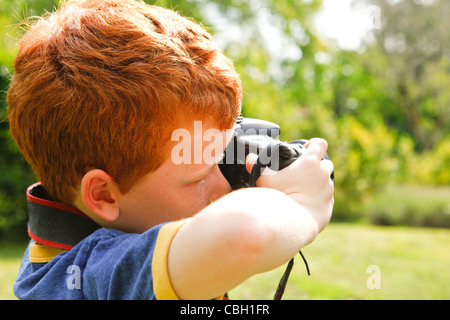 Un jeune garçon, âgé de 7 ans, en utilisant un appareil photo reflex numérique dans un jardin ensoleillé. Banque D'Images