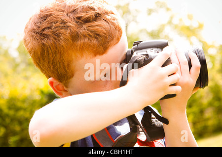 Un jeune garçon, âgé de 7 ans, en utilisant un appareil photo reflex numérique dans un jardin ensoleillé. Banque D'Images