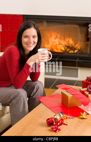 Happy woman wrapping Christmas present par cheminée bénéficiant d'une boisson chaude Banque D'Images