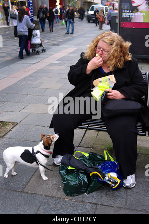Femme assise sur un banc public dans la rue Sauchiehall Street, Glasgow en train de manger un sandwich avec son petit chien à la recherche sur. L'Écosse, Royaume-Uni, Banque D'Images