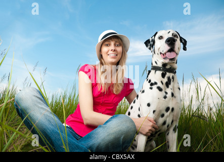 Belle jeune femme avec chapeau assis dans l'herbe avec son chien dalmatien animal et souriant. À la fois dans l'appareil. Ciel bleu Banque D'Images