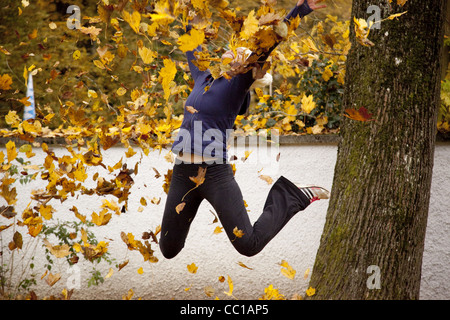 Une fille saute avec les jambes pliées et jette des feuilles jaunes dans l'air sur un après-midi d'automne. Banque D'Images