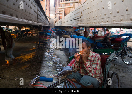 Sans-abri d'un conducteur de pousse-pousse cycle fume sous un pont sur un parking à côté de la rivière Yamuna où il dort. New Delhi Banque D'Images