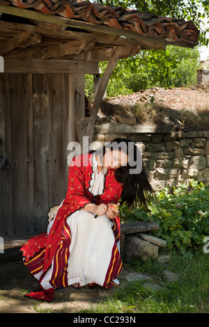 Belle jeune femme en costume national bulgare peeling une pomme dans le vieux village de Jeravna Banque D'Images