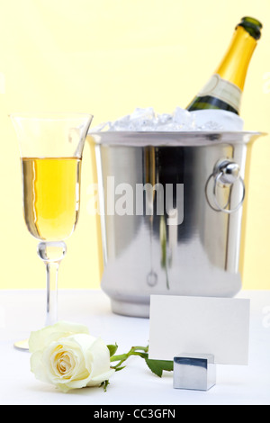 Photo d'une rose blanche sur une table avec nappe blanche et un verre de champagne et une bouteille dans un seau à glace à l'arrière-plan. Banque D'Images