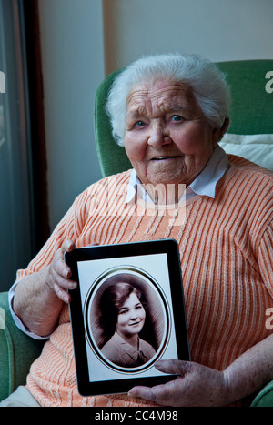 98 ans vieille dame tenant un ordinateur tablette iPad, affichant un portrait sépia d'elle-même pris il y a 80 ans, à l'âge de 18 ans Banque D'Images