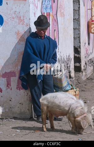 Un homme d'âge moyen dans l'habillement traditionnel équatorien s'appuie contre le coin d'un bâtiment avec un cochon en laisse en face de lui. Banque D'Images