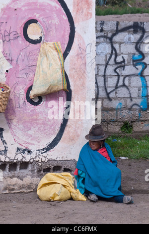 Une très vieille femme indienne andine est assis sur le sol près d'un mur de graffiti marqué Saquisili, Equateur, Amérique du Sud. Banque D'Images