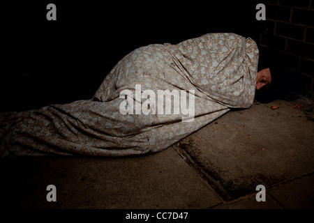 Image représentant un sans-abri couvert à dormir dans une couverture sur la chaussée de pierre froide. Banque D'Images