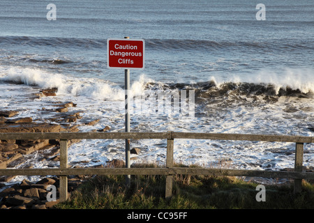 Panneau 'Attention' avec des falaises dangereuses vagues dans l'arrière-plan, Seaton Sluice, North East England UK Banque D'Images