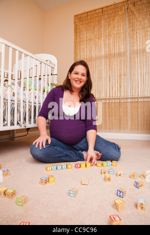 Femme enceinte avec des blocs de construction, assis sur le plancher à côté de lit-bébé Banque D'Images