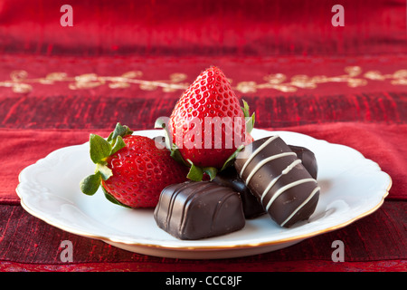 Le chocolat noir bon-bons et des fraises sur une fine plaque blanche avec décoration d'or de la Chine sur la jante. Fond texturé rouge. Banque D'Images