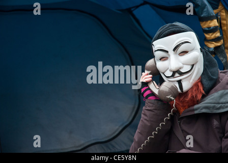 Un manifestant masqué portant un masque de Guy Fawkes, campé à St pauls occupy london site sur un vieux téléphone, tente dans l'arrière-plan. Banque D'Images