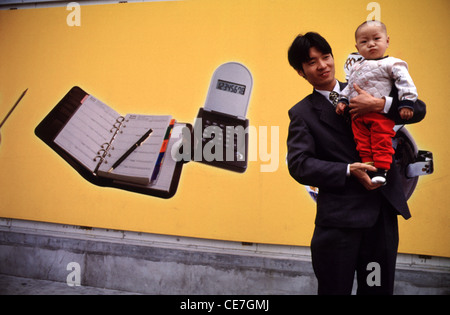 Un jeune homme chinois en costume noir tient son fils en face d'une affiche présentant du matériel de bureau en Chine Banque D'Images