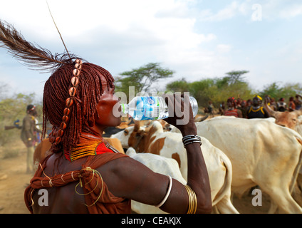 Hamer femme habillée traditionnellement chez les bovins de boire de l'eau bouteille plastique l'Ethiopie Banque D'Images