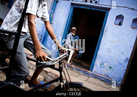 Un garçon portant des sandales bleu chevauche son vélo passé où un homme assis dans un bleu blue house à Agra, en Inde. Banque D'Images