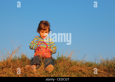 Petite fille en t-shirt à rayures avec sac rouge est assis sur l'herbe sèche Banque D'Images