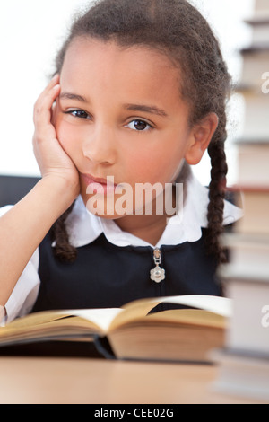 Une belle young African American girl la lecture dans une salle de classe avec une pile de livres en face d'elle Banque D'Images