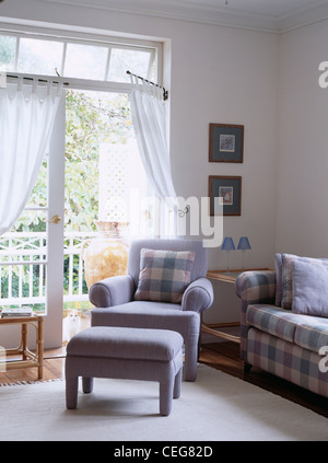 Fauteuil et repose-pieds bleu en face de portes-fenêtres avec rideaux de voile blanc sur les tiges de portiere Banque D'Images