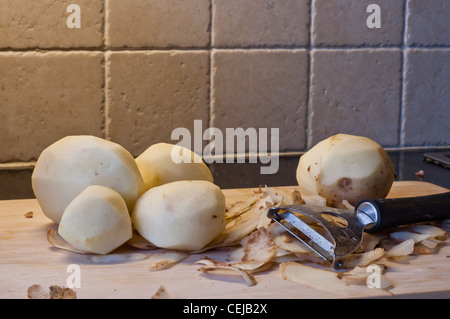 Les pommes de terre sur une planche après avoir été pelée et une pomme de terre pelées partiellement entouré par des peelings et le métal éplucheuse de pommes de terre. Banque D'Images