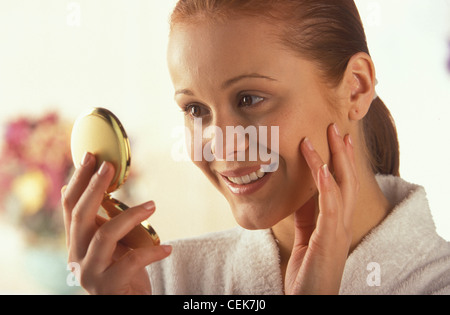 Femelle alezan cheveux visage portant un peignoir blanc à la recherche d'elle-même dans mirrtouching compact joue doigts smiling Banque D'Images