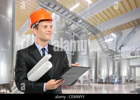 Un travailleur de la construction avec helmet holding blueprints et écrit sur un presse-papiers dans une usine Banque D'Images