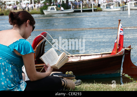 Une jeune femme assise sur la berge de la lecture d'un livre à l'assemblée annuelle de l'Henley Royal Regatta. Banque D'Images