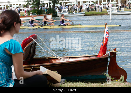 Une jeune femme assise sur la rive à la recherche à partir d'un répertoire pour regarder une vitesse de course passé à l'assemblée annuelle Henley Royal Regatta. Banque D'Images