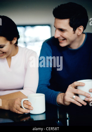 Cheveux bruns courts MMale portant haut bleu et noir et anneau argent sitting at table holding white mug sourire montrant des dents, Banque D'Images