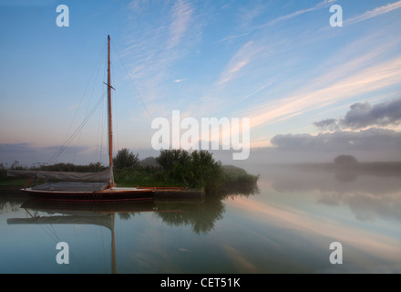 Un bateau à voile dans le brouillard de l'aube sur la rivière Thurne dans les Norfolk Broads. Banque D'Images