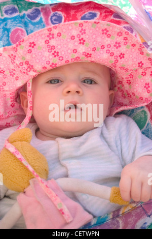 Baby wearing pink floral chapeau de soleil et babygro rayé bleu et blanc à motifs multicolores, assis dans la pram holding teddy Banque D'Images