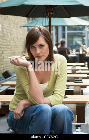 Longueur de l'épaule droit femelle de cheveux brune, porter un jean bleu, vert cardigan, assis sur un banc, les genoux, les coudes reposant Banque D'Images