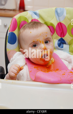 Un bébé de sexe féminin, les cheveux juste assis dans une chaise haute, le port d'un carré de mousseline rose et bib en plastique, manger en purée de couleur orange Banque D'Images