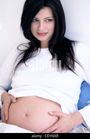 Une femme enceinte de long cheveux brune portant des T-shirt à manches longues blanc assis appuyé contre l'oreiller, les mains Banque D'Images