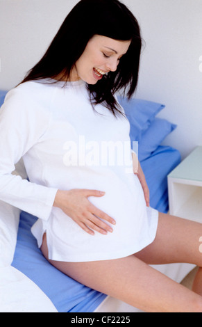 Profil de semi long femelle enceinte cheveux brunette wearing white top assis sur le bord du lit à l'estomac holding Banque D'Images