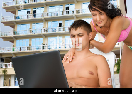 Jolie femme et jeune homme couchée sur des chaises longues sur la plage près de l'hôtel, l'homme à la recherche dans un écran d'ordinateur portable Banque D'Images