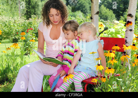 Jeune femme lit le livre à deux petites filles en jardin Banque D'Images