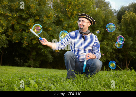 L'homme en costume pirate joyeux est assis sur l'herbe et en soufflant des bulles de savon. Banque D'Images