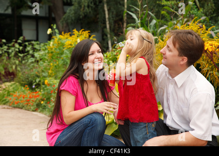 Les parents observent que leur fille en robe rouge souffle des bulles de savon dans le jardin d'été. Banque D'Images