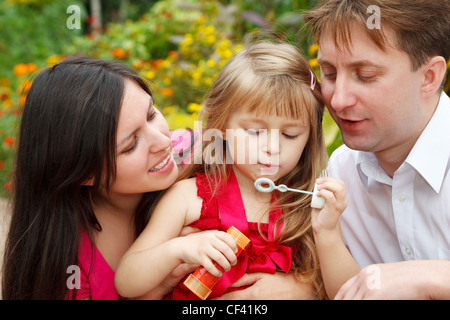 Les parents observent que leur fille en robe rouge souffle des bulles de savon dans le jardin d'été. Close up. Banque D'Images