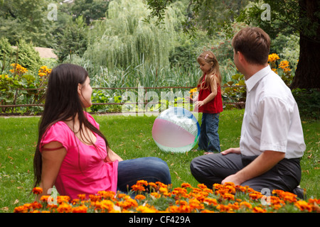 Petite fille joue avec grand ballon gonflable dans le parc en après-midi. Observer les parents de celui-ci assis sur la pelouse. Banque D'Images